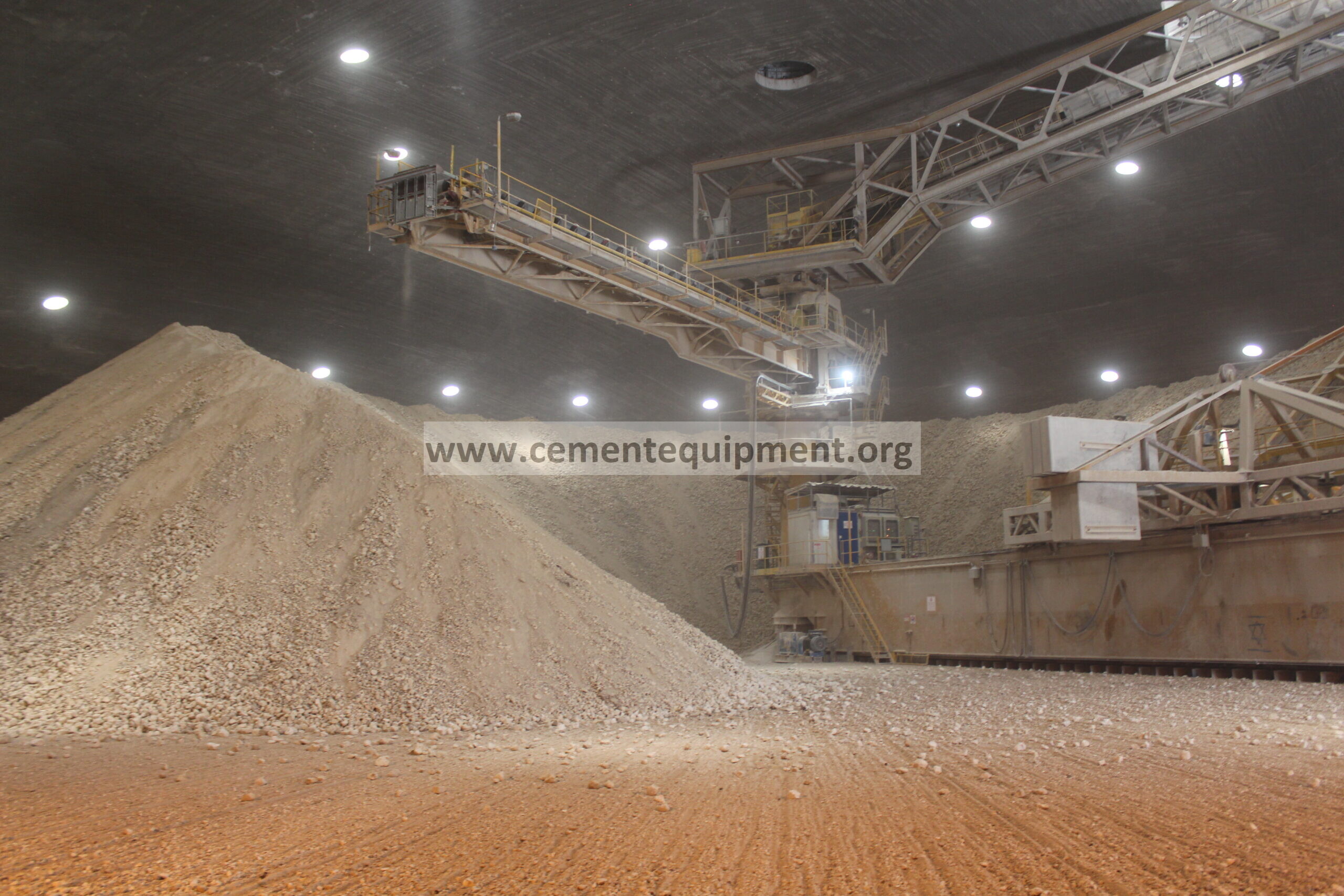 Covered bulk-material stockyard with overhead conveyor and material stockpiles; slew-drive mechanism not visible